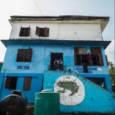 Photo of 3-storey blue Liberian school with two students in doorway