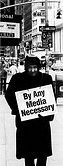 Man on sidewalk holding sign reading "By Any Media Necessary"