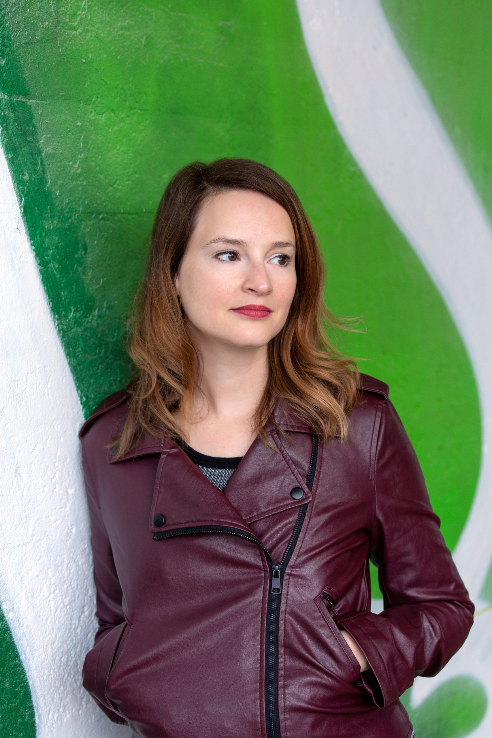 Person with shoulder-length dark hair, wearing a leather jacket, leaning against a wall and looking away from the camera