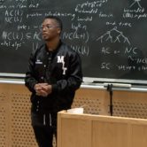 Lupe Fiasco presenting in front of a lecturn and wearing a black jacket with the letters MIT on it in white